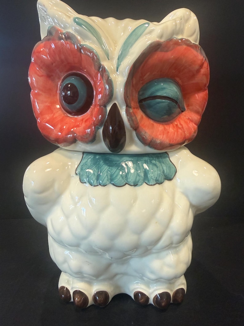 May include: A ceramic owl-shaped cookie jar. The owl has a white body, red and teal eyes, and a teal collar. The beak is brown. The jar is a decorative kitchen item.