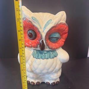 May include: A ceramic owl figurine, approximately 12 inches tall, with a winking expression. The owl features a cream-colored body, red and blue accents on the face, and brown feet. It has a blue ruffled collar and is standing upright.