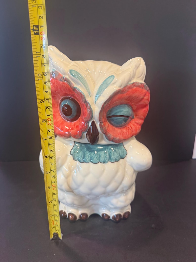 May include: A ceramic owl figurine, approximately 12 inches tall, with a winking expression. The owl features a cream-colored body, red and blue accents on the face, and brown feet. It has a blue ruffled collar and is standing upright.