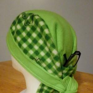 Puede incluir: Sombrero de forro polar verde con un patrón a cuadros verde y blanco y bordado de trébol. El sombrero está atado en la parte posterior y tiene un bolsillo para gafas.