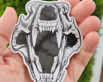 May include: A black and white sticker featuring a detailed illustration of a bear skull with open jaws. The sticker has the text "Kodiak, Alaska" at the bottom. The artist's signature is visible on the right side.