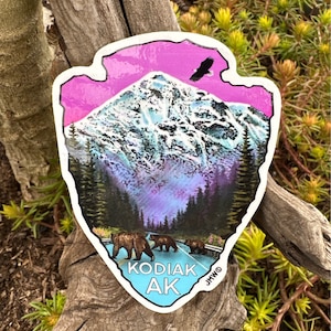 May include: A sticker with a mountain scene, an eagle, and bears crossing a road. The sticker has a pink and blue sky, a snowy mountain, and the text "KODIAK AK". The sticker is in the shape of an arrowhead.
