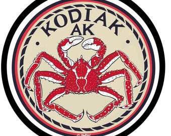 May include: A round sticker featuring a red crab with white accents, set against a beige background. The text "KODIAK AK" is arched above the crab. The sticker has a black, white, and red border.