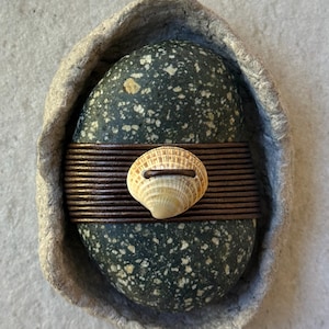 May include: A handcrafted decorative piece featuring a dark speckled stone wrapped with a copper-colored band and a small seashell. The stone is set within a gray, textured, paper-like frame, creating a unique, natural aesthetic.