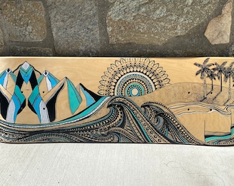 May include: A wooden skateboard deck with a hand-painted design featuring a mountain range, a mandala sun, waves, and palm trees. The design is done in black, blue, green, and gold paint.