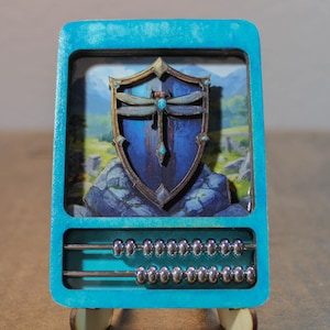 Puede incluir: Un ábaco de color turquesa con un diseño de escudo que presenta una libélula y un paisaje. El ábaco tiene dos filas de cuentas y está montado sobre pequeños pies de madera. El escudo es azul y dorado.