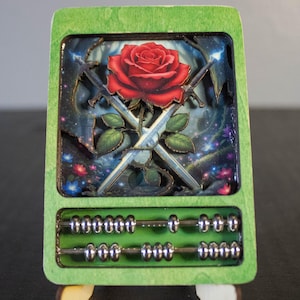 Puede incluir: Un ábaco de madera verde con un diseño recortado detallado. El diseño presenta una rosa roja, espadas cruzadas y un fondo estrellado. El ábaco tiene cuentas plateadas y una forma rectangular.