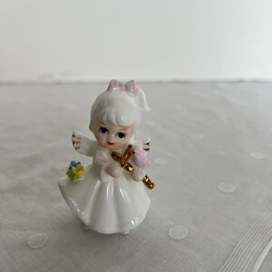 May include: A white porcelain figurine of an angel girl with blonde hair, a pink bow, and a pink bird in her hand. She is wearing a white dress with gold wings and flowers.