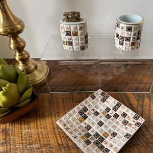 May include: Two small ceramic containers with a brown and white checkered pattern, one with a gold top and a lighter, on a clear acrylic shelf. A brown and white checkered tile coaster is in the foreground.