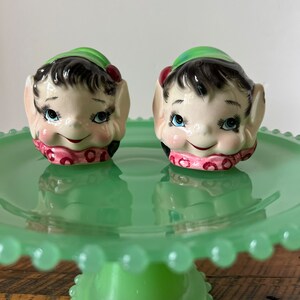 Peut inclure: Une paire de salières et poivrières en céramique en forme d'elfes avec des chapeaux verts et des écharpes roses. Les salières sont posées sur un support à gâteau en céramique vert.