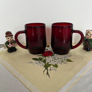Könnte beinhalten: Zwei rote Glasbecher mit Henkeln. Die Becher stehen auf einer weißen Tischdecke mit Blumenmuster.