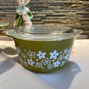 以下が含まれることがあります： 白い花柄の緑色のキャセロール皿と透明なガラスの蓋。皿は白いテーブルクロスの上に置かれています