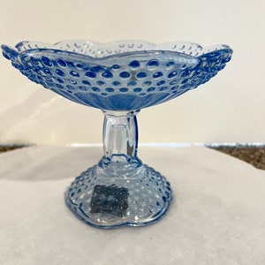 May include: A light blue glass pedestal bowl with a scalloped rim and a textured, hobnail design. The bowl sits on a clear, stemmed base. The bowl is likely used for serving desserts or displaying decorative items.