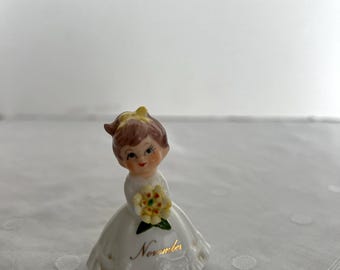 May include: A white porcelain figurine of a girl wearing a white dress and holding a yellow flower bouquet. The figurine is inscribed with the word "November".