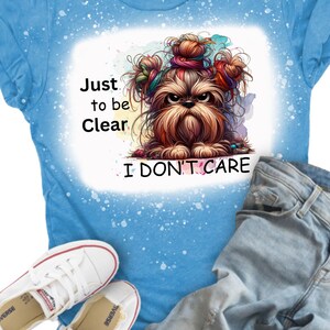 Puede incluir: Una camiseta azul con un dise&ntilde;o de salpicaduras blancas. La camiseta presenta una imagen de dibujos animados de un perro con lazos de pelo coloridos y el texto "Just to be Clear I Don't Care".