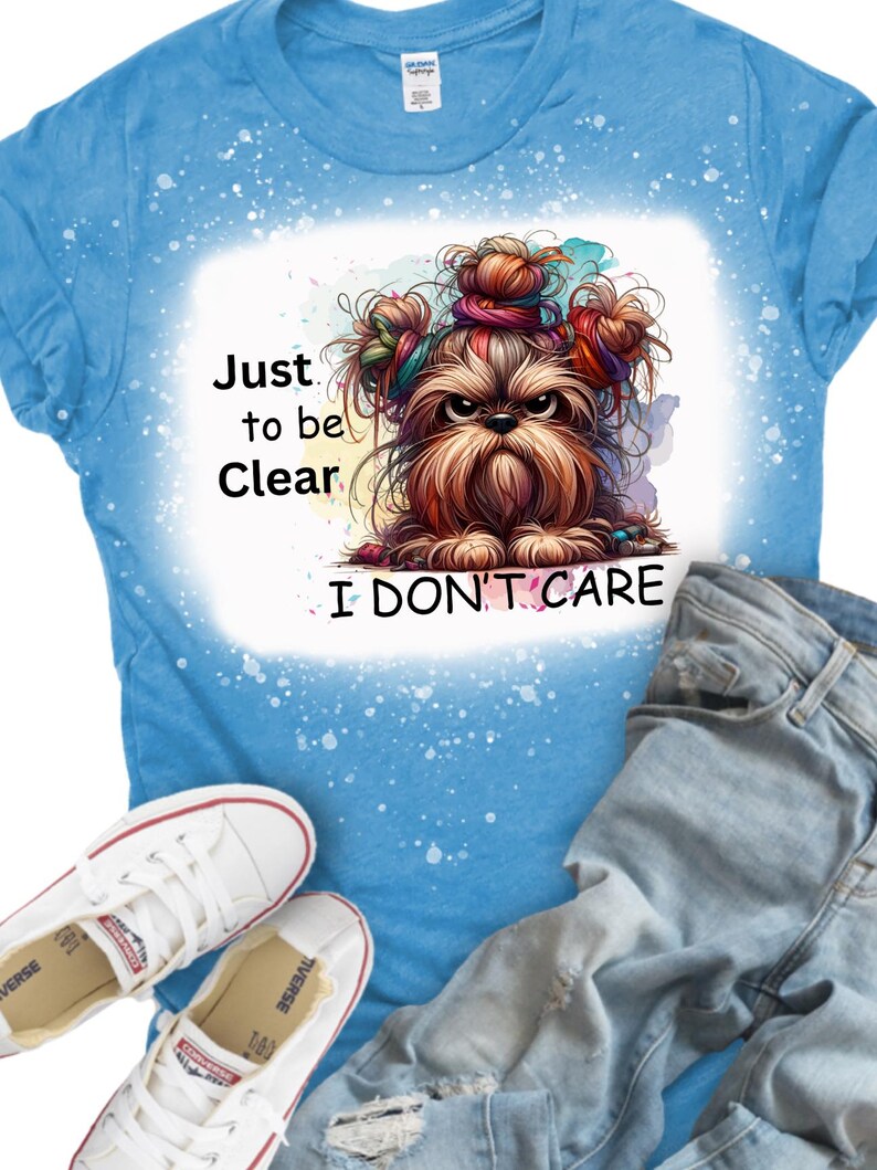 Puede incluir: Una camiseta azul con un dise&ntilde;o de salpicaduras blancas. La camiseta presenta una imagen de dibujos animados de un perro con lazos de pelo coloridos y el texto "Just to be Clear I Don't Care".