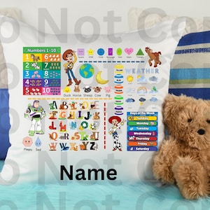 Op de afbeelding: Witte kussensloop met educatieve afbeeldingen, waaronder cijfers, letters en weersymbolen. Bevat stripfiguren en de tekst "Name". Een teddybeer en kussens zijn op de achtergrond.