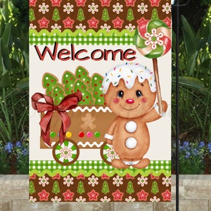 May include: A festive garden flag featuring a gingerbread man holding a lollipop. The flag has a "Welcome" message and a gingerbread house design with Christmas tree cookies. The border has a brown and green pattern with gingerbread men and Christmas trees.