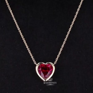 May include: A gold-colored necklace with a heart-shaped, deep red gemstone pendant. The pendant is set in a gold-colored bezel and hangs from a delicate chain. The background is black.