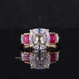 Könnte beinhalten: Ein goldener Ring mit einem großen, quadratischen, klaren Edelstein, umgeben von drei kleineren, rechteckigen, roten Edelsteinen.