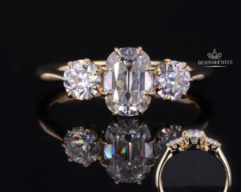 May include: A gold ring with three clear, sparkling gemstones. The center stone is oval-shaped, flanked by two round stones. The ring is set against a black background, with a reflection of the ring visible below. The text "D/SOSMUEWELS" is in the upper right corner.