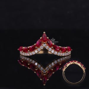 May include: A gold-toned ring with a crown design, featuring marquise-cut red gemstones and small white gemstones. The ring is displayed on a reflective black surface, with a side view of the ring also shown.