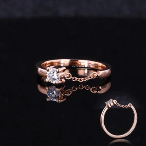 May include: A rose gold ring featuring a single diamond and a delicate chain attached. The chain hangs down from the ring and is visible in the reflection of the ring.