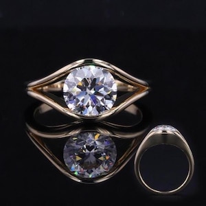 May include: A gold ring featuring a large, round, brilliant-cut clear gemstone set in a tension-style mounting. The band splits into two smooth, polished gold arms that curve around the center stone, creating an open, bypass-like silhouette.