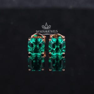 May include: A pair of emerald-cut, vivid green gemstone stud earrings. The earrings are set in a rose gold metal, with the brand name "DASOSMJEWELS" visible. The gemstones are square-shaped and reflect light.