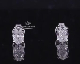 May include: A pair of oval-cut diamond stud earrings. The diamonds are set in a silver metal, and the earrings are displayed against a black background. The earrings are reflecting on the surface.