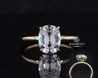 May include: A gold ring with a large, clear, cushion-cut gemstone. The ring is set with four prongs and has a thin band. The ring is displayed on a reflective black surface. The text "DASOSMUEWELS" is visible.