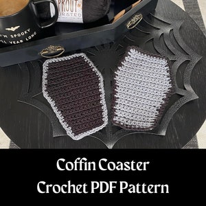 May include: Two crocheted coffin-shaped coasters, one black with a gray trim and one gray with a black trim, on a black surface with a spiderweb design.