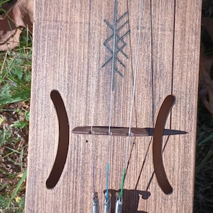 May include: A wooden musical instrument with strings and a carved design. The instrument has a brown wooden body with a dark brown wood trim. The strings are attached to tuning pegs at the top of the instrument and run down to a bridge at the bottom. The instrument has a carved design on the front, which appears to be a stylized tree or plant.