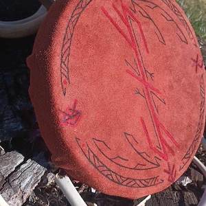 Könnte beinhalten: Eine rote Trommel aus Wildleder mit schwarzen und roten Designs. Die Trommel liegt auf einer Holzoberfläche mit weißen Geweihstangen.