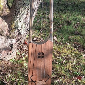 Puede incluir: Una lira de madera hecha a mano con cuerdas, con una caja de resonancia marrón oscuro y un marco marrón más claro. El instrumento tiene un diseño decorativo de cuatro pétalos y se encuentra en un entorno natural al aire libre.