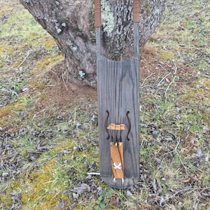 May include: A handmade wooden instrument with a dark gray finish, featuring brown leather-wrapped handles and decorative elements. The instrument is suspended against a tree trunk, showcasing its unique design and craftsmanship.