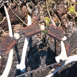 Puede incluir: Un cordón de cuero marrón con cuentas de madera y dijes. Los dijes son rectangulares y tienen intrincados diseños tallados en ellos. El cordón está envuelto alrededor de un par de cuernos de ciervo blancos.