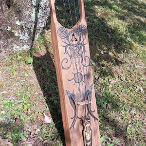 May include: A wooden lyre with five strings and intricate carvings. The lyre is resting on a bed of green grass and brown leaves.