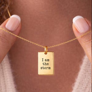 May include: Gold-colored necklace with a rectangular pendant. The pendant has the phrase "I am the storm" engraved in a simple font. The necklace is held by a person.