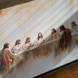 May include: A framed canvas print of the Last Supper. The image shows figures in robes seated at a long table. The colour palette includes warm tones of brown, beige, and white, with a wooden frame.