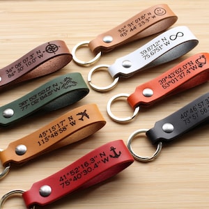 May include: Seven leather keychains with different colored leather and engraved coordinates, symbols, and text. The keychains are on a wooden surface.