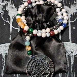 以下が含まれることがあります： 円形のデザインが施された銀製のペンダントが付いた白と黒のビーズネックレス。ネックレスは黒いベルベットのポーチに置かれています。