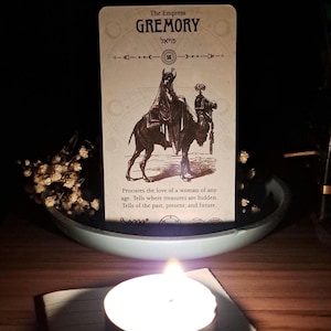 以下が含まれることがあります： 「The Empress Gremory」と書かれたタロットカードのイラスト。ラクダに乗った人物が描かれています。カードには英語とヘブライ語のテキストがあります。明るい青色のボウルの中にあり、手前にろうそくが灯っています。