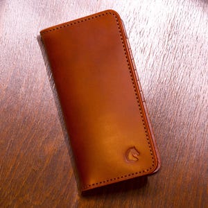 Könnte beinhalten: Eine braune Ledergeldbörse mit einer genähten Kante und einem Pferdekopf-Logo. Die Geldbörse ist rechteckig und geschlossen. Das Leder hat eine glatte, polierte Oberfläche. Die Geldbörse liegt auf einer Holzoberfläche.