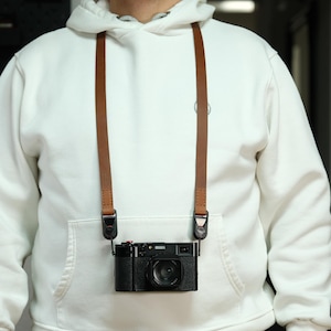 Könnte beinhalten: Eine schwarze Kamera mit einem braunen Lederriemen hängt am Hals einer Person, die einen weißen Kapuzenpullover trägt. Die Kamera hat ein Objektiv und mehrere Knöpfe. Der Lederriemen ist mit Metallclips an der Kamera befestigt.