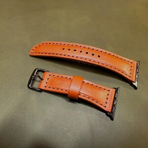 Könnte beinhalten: Zweiteiliges Uhrenarmband aus hellbraunem Leder mit schwarzen Nähten. Das Armband hat eine schwarze Schnalle und Anschlüsse. Das Leder-Uhrenarmband wird auf einer grünen Oberfläche gezeigt.