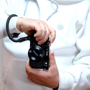 Könnte beinhalten: Eine schwarze Digitalkamera mit Objektivdeckel mit der Aufschrift "SpeedHunt" wird gehalten. Die Kamera hat eine schwarze Handschlaufe mit roten Nähten. Die Person trägt einen weißen Kapuzenpullover.