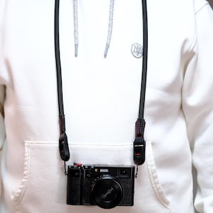 Könnte beinhalten: Eine schwarze Kamera mit Objektivdeckel und schwarzem Gurt hängt um den Hals einer Person, die einen weißen Kapuzenpullover trägt. Der Kameragurt ist schwarz mit roten Nähten. Der Kapuzenpullover hat eine Vordertasche.