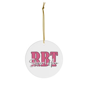 May include: A white ceramic ornament with the text "RBT Super Hero" in pink and black. The ornament is round and has a gold string for hanging. The text is in a bold, blocky font.