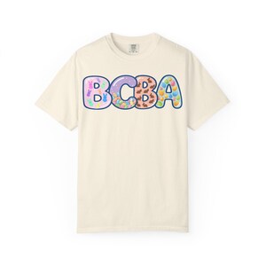 以下が含まれることがあります： クリーム色のTシャツ。大きなカラフルなバブル文字で「BCBA」と書かれています。文字はウサギや卵など、イースターをテーマにしたグラフィックで埋められています。シャツはクラシックなクルーネックと半袖です。